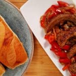 Sausage Peppers and Onions on a white plate. hoagie rolls on blue plate. bamboo cutting board