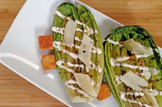 Grilled Caesar Salad drizzled with dressing, croutons, shaved parmesan cheese on white plate on bamboo cutting board