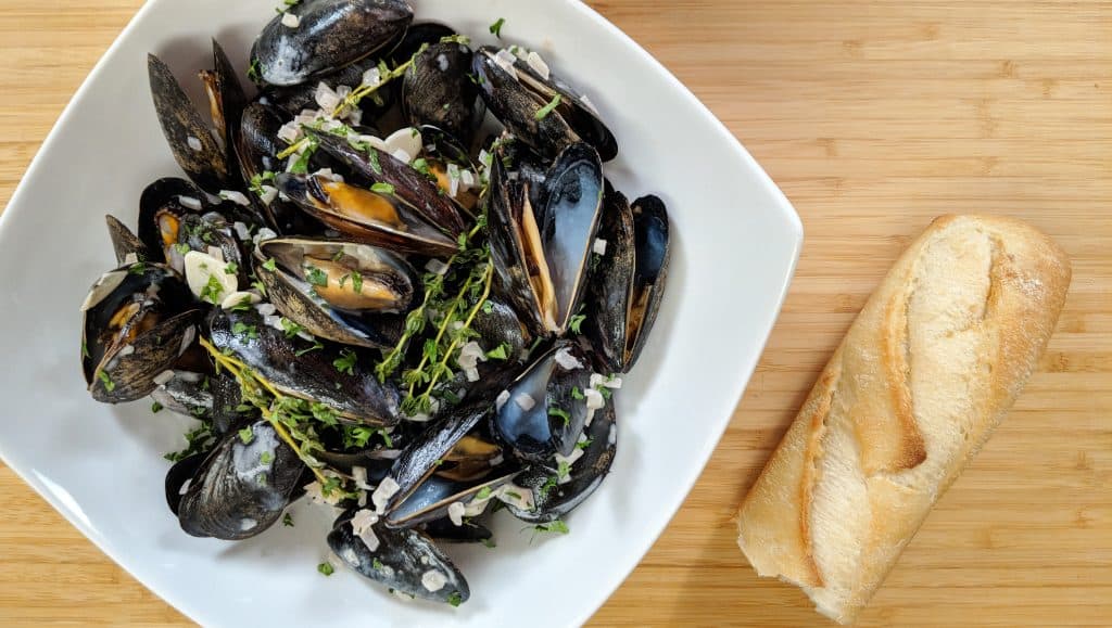 A bowl of black mussels with a crusty baguette. Garlic, shallots and thyme flavor the dish.