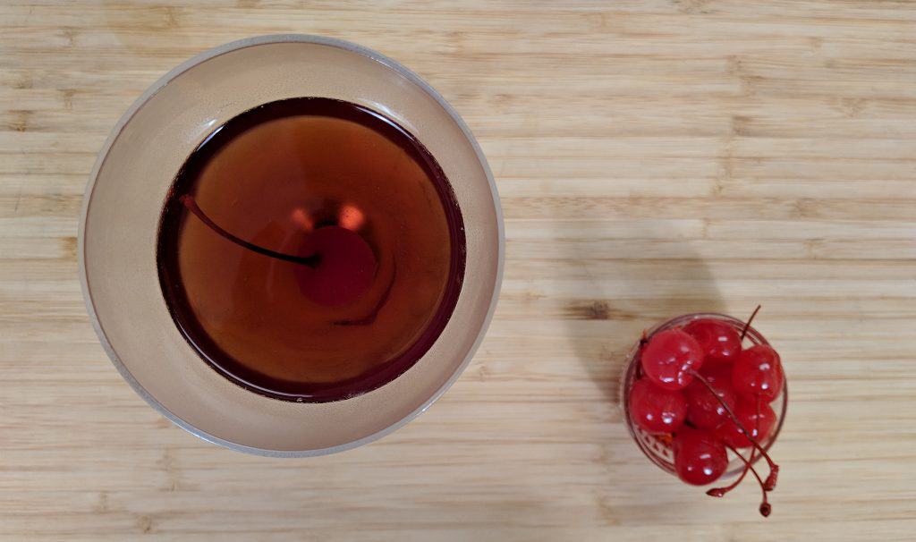 Ovehead view of manhattan cocktail in nick and nora glass with maraschino cherries on a bamboo cutting board