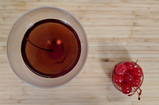 Ovehead view of manhattan cocktail in nick and nora glass with maraschino cherries on a bamboo cutting board