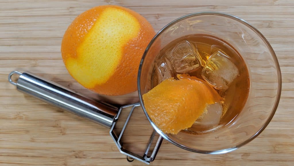 Overhead View of Old Fashioned Cocktail with Vegetable Peeler and Peel Orange and Orange Rind on a Bamboo Cutting Board