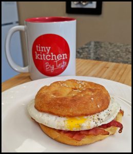 Keto Bagel Egg Sandwich with Bacon on a white plate with a Tiny Kitchen Big Taste Coffee Mug